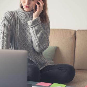 En kvinna klädd i en grå stickad tröja som sitter i en soffa och pratar i telefon. Hon tittar mot en bärbar dator som syns delvis i bilden. Bakgrunden är ljus och avskalad.